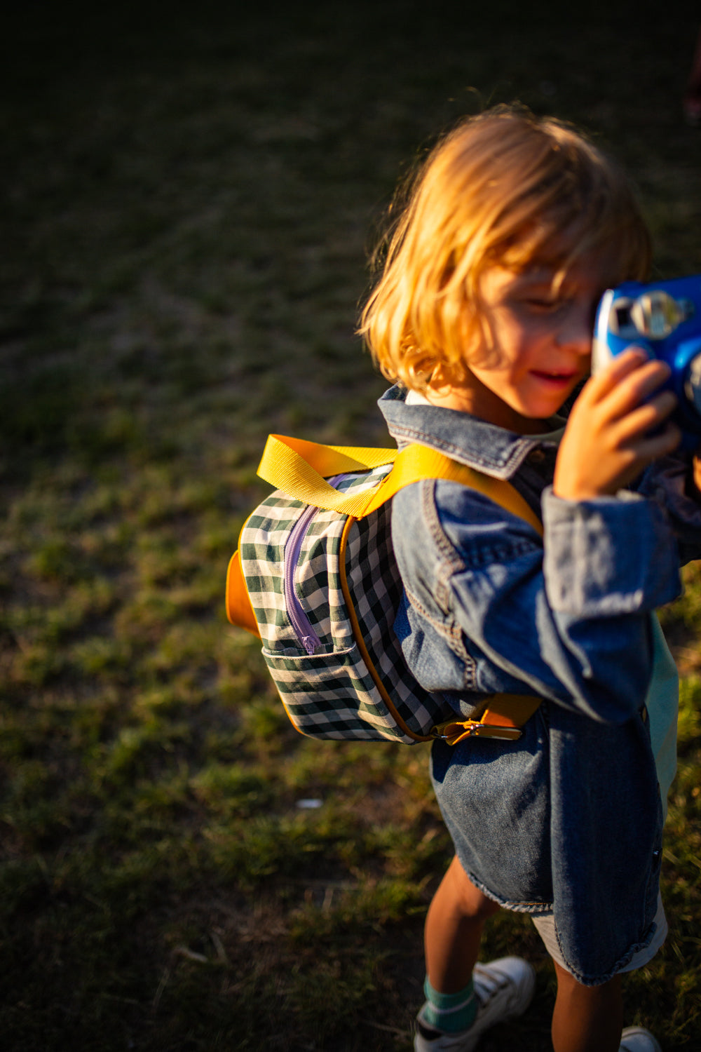 a little boy with a small gingham backpack pretends to take pictures with a toy camera. small checkered backpack