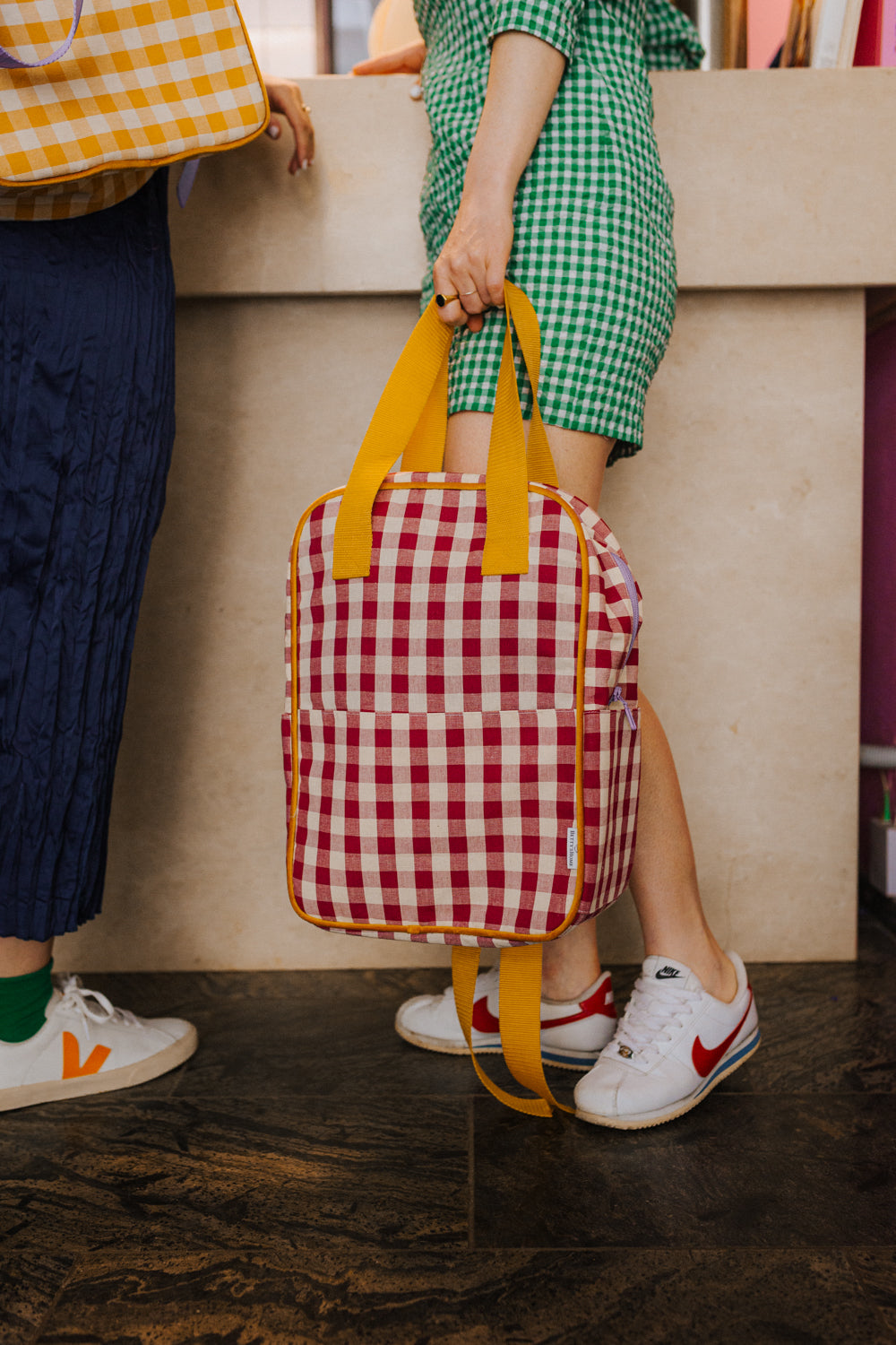 woman in green dress standing in restaurant with red checkered backpack in hand. Red Gingham Backpack by bettys home