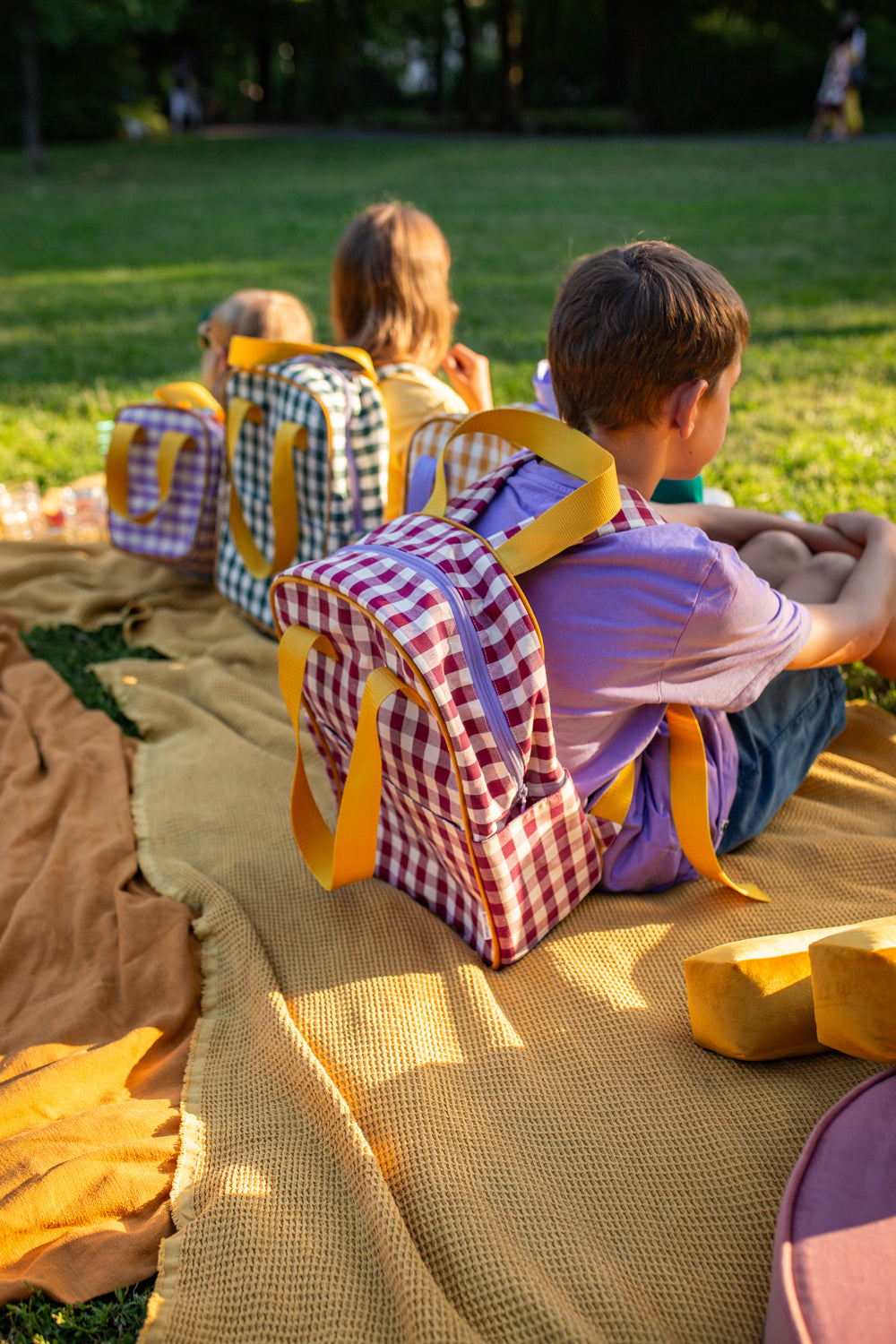 little kids sitting on blanket during picnic with green checkered backpack and red checkered backpack. gingham backpacks