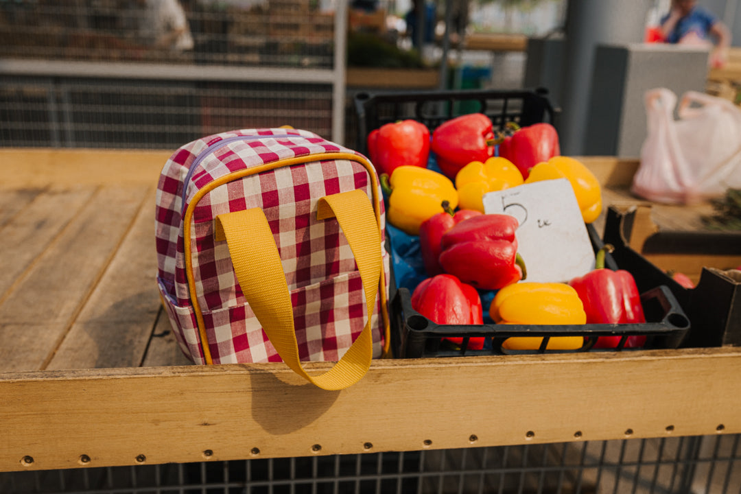 small red gingham backpack stands on the counter by the basket of peppers. checkered small backpack 