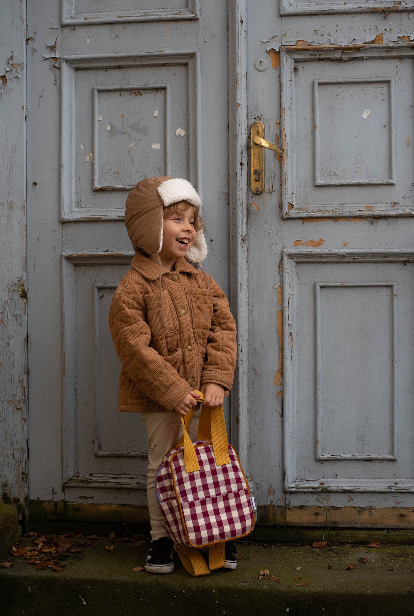 cute little boy with small red checkered backpack standing stands against the backdrop of an old door 