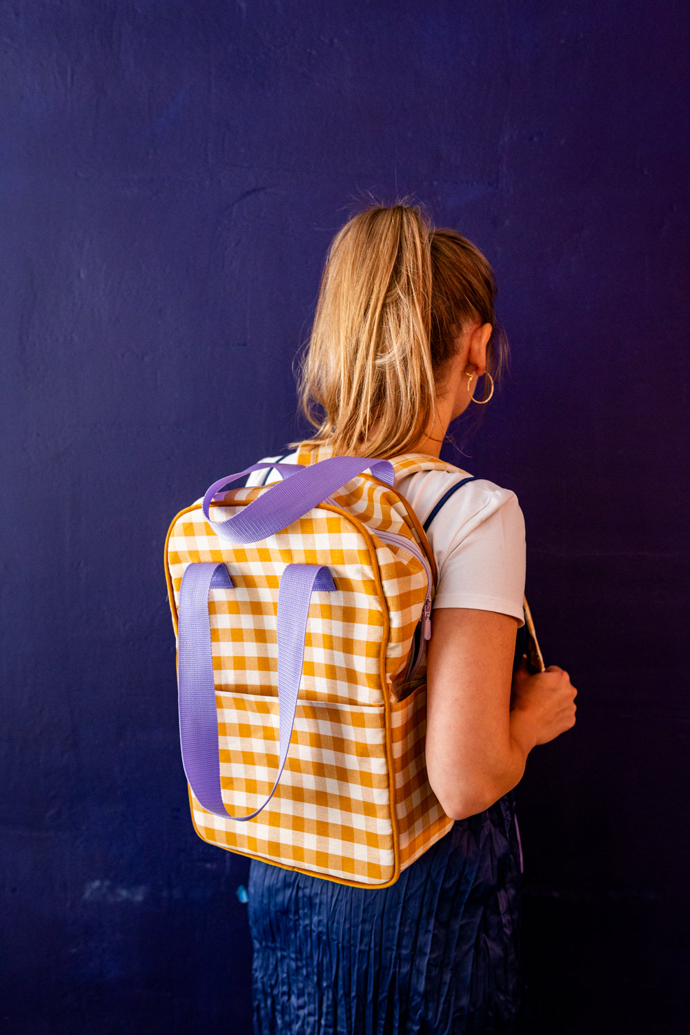 young woman in denim skirt stands against blue wall with yellow gingham backpack by bettys home. Checkered backpack in yellow