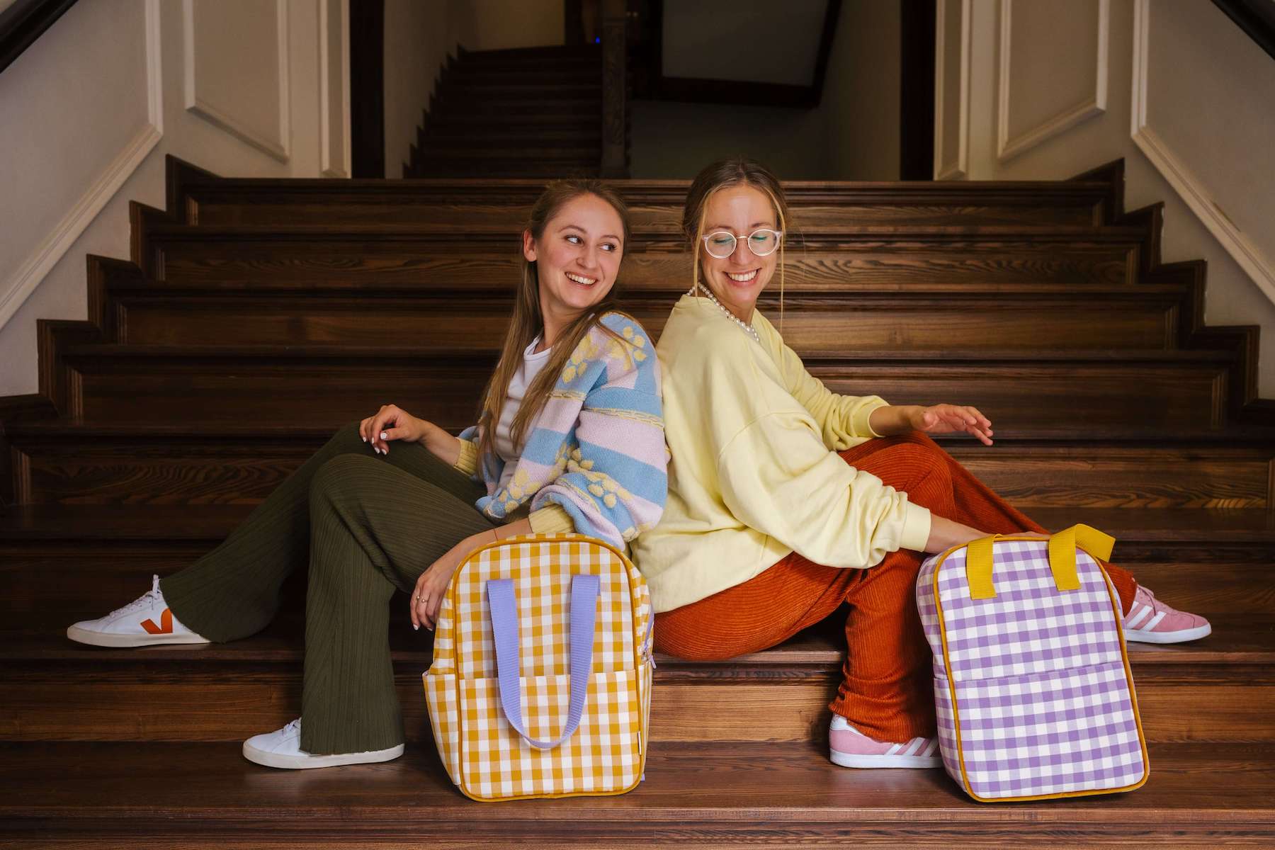 two friends sitting on stairs and smiling. At front there are gingham backpacks by bettys home