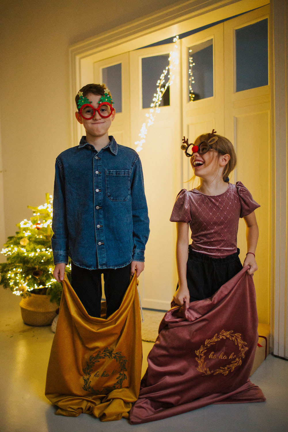 a boy and a girl dressed for Christmas stand next to a Christmas tree with Christmas gift bags by bettys home