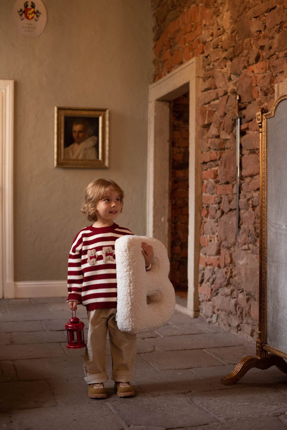 little boy standing in hall in red stripe sweater with boucle letter cushion B in hand