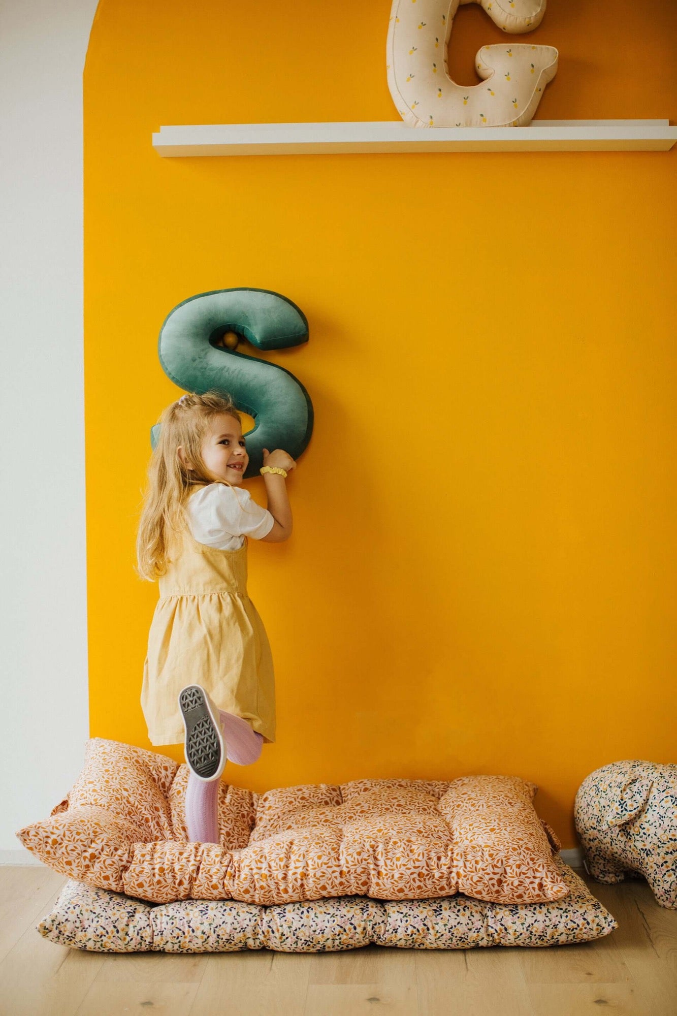 little girl standing on floor mats with velvet letter cushion s mint in hands on yellow wall
