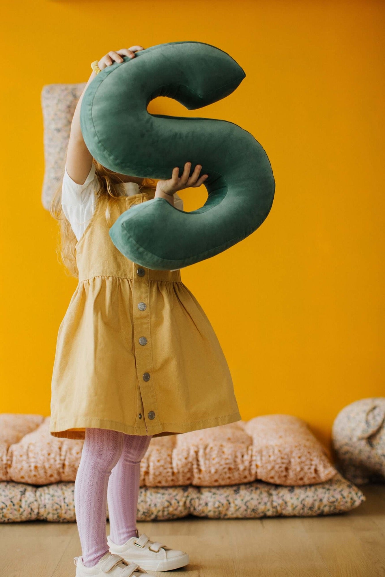 little girl with velvet letter cushions s mint in here hands standing on yellow wall. 
