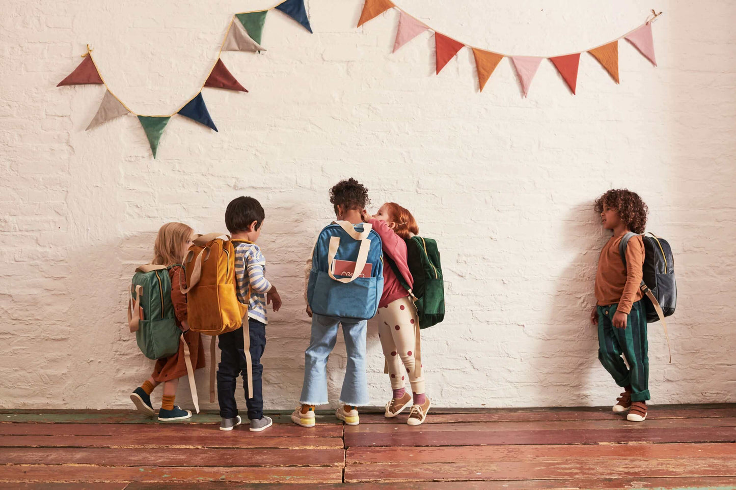Children standing in school at recess with colorful velour backpacks from Bettys Home. Above the children hangs a wall decoration in the form of a garland of triangles from Bettys Home 